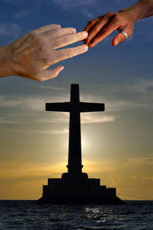 Large concrete cross silhouette over a marine sunset with a Caucasian and a black hand touching on top in unity.の写真素材
