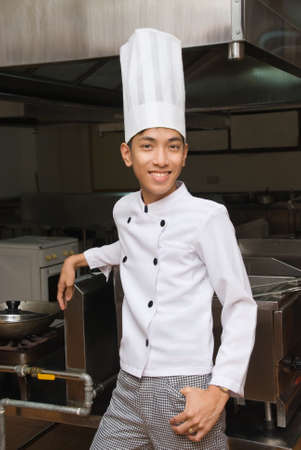 Young smiling Chinese oriental manager, chef or cook in a restaurant kitchen, proud and leaning back in relaxed way against the cooking equipment.の写真素材