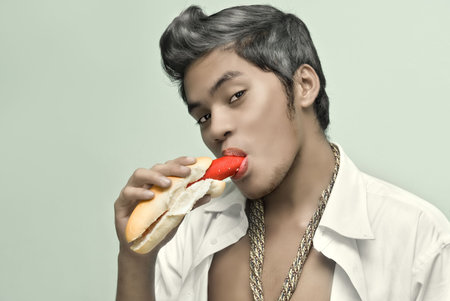 Portrait of young Asian man with smoothened fantasy skin slowly sucking and eating hot dog in shirt...