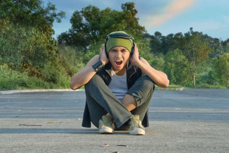 Young Hispanic rapper dude in casual grunge clothing with cap and headphone sitting in the middle of a green suburban street covering his ears for sound or noise overload, shouting with an intense expression of disgust on his face.の写真素材