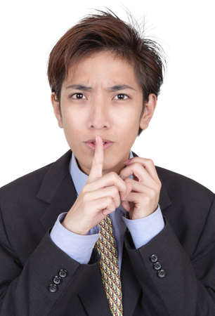 Portrait of young Chinese businessman hushing with fingers on lips and a mesmerizing urging gaze insisting on discretion about a deal. Isolated over white.の写真素材