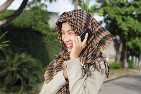Attractively laughing happy Asian Muslim girl with headscarf or hijab and in formal jacket calling by cell phone outside on a tropical affluent residential street.の写真素材