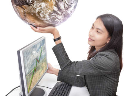 Happy young Asian businesswoman in corporate outfit sitting at her desk in front of her computer screen with spreadsheet data and holding up a globe. Isolated over white. Globe courtesy NASA/JPL.の写真素材