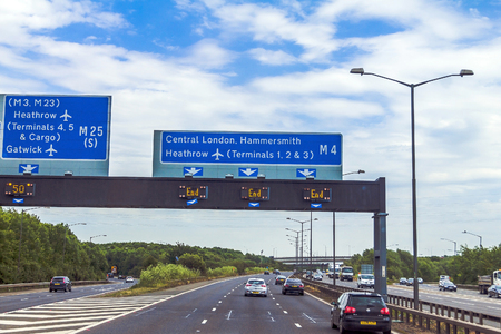 Intensive left-hand traffic on British four lane motorway M4 between Windsor and London  with active electronic overhead information sign at grey cloudy  summer day. UKのeditorial素材