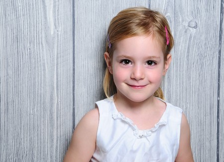 Portrait of a cute four-year smiling blonde girl in white dress and colorful barrettes in her hair standing on paper gray  wooden fence backgroundの写真素材