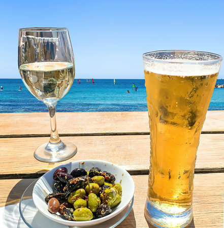 Wooden table with light beer in the mug , white wine in the glass and olives in white plate at the beach on blue sea and sky background.の写真素材