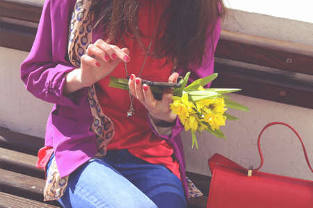 Stylish girl in blue jeans with bouquet of yellow daffodils flowers sitting on the bench. Business lady with phone on the street. Fashionable brunette woman with cell in hands.の写真素材