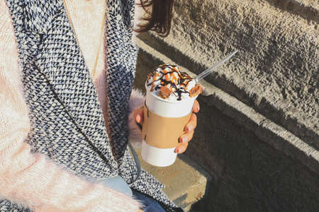 Stylish fashionable woman in light pink sweater holding in hand paper cup with tasty sweet coffee with cream, chocolate and caramel. Vogue woman drinking coffee in the city.の写真素材