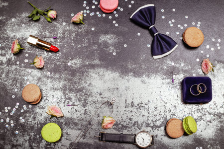 Happy wedding day concept. White gold luxury necklace, macaroones, two wedding rings on the velvet box, deep blue bowtie, roses, lipstick and men's watch on the table. Place for text.の写真素材
