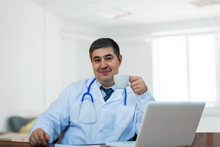 A male doctor with a cup of coffee at the computer. He looks at the camera and smiles. Coffee break.