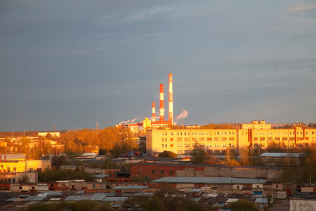 The image shows a thermal power plant with tall chimneys illuminated by the rays of the setting sun against the sky. The scene is ideal for content related to thermal energy and heating plants.の写真素材