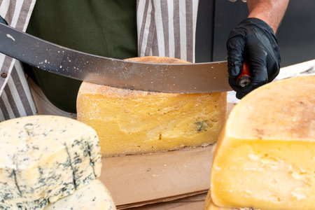 A vendor slices a piece of cheese with a knife at a farmers market. Close-up of hands cutting a...