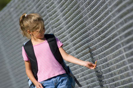 Little girl walking to school by school fence on beautiful sunny day wth back pack and pencilの写真素材