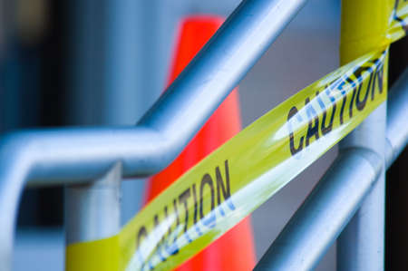 Yellow caution tape on stair rail with orange traffic cone behind, very shallow depth of field.の写真素材