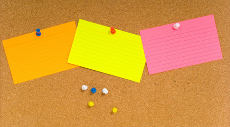 Bright, blank neon colored notecards on brown corkboard or bulletin board with plastic pushpins,...