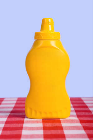 A yellow squeeze bottle in fornt of a blue sky on a  red and white checked or gingham tablecloth with no lableの写真素材
