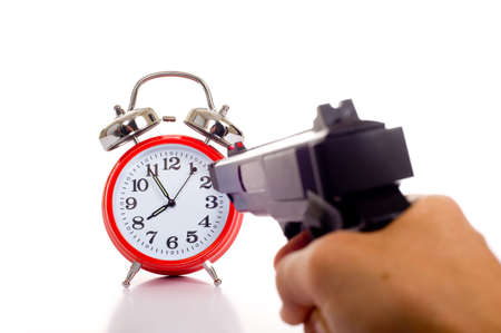A vintage red alarm clock with a gun pointing at it on a white background.  Conceptual image of frustration with time or killing time etc.の写真素材