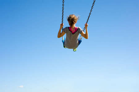 A young girl playing on a swingset in front of a blue sky.  Girl is swinging very high in swing, with copy spaceの写真素材