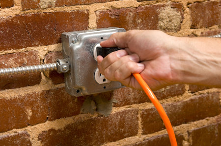 a man's hand plugging in an extension cord in  a junction box electrical outlet on a brick wall,...