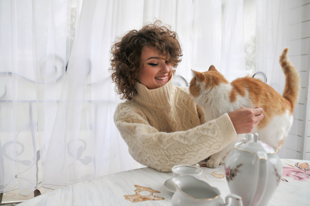 Happy woman in warm sweater with curly hair, smiling and cuddling with red catの写真素材
