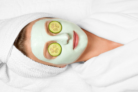 Beautiful young woman is getting facial clay mask at spa, lying with cucumbers on eyesの写真素材