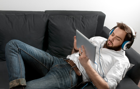 guy with a beard in a white shirt and headphones listens music,Young and smiling man sitting on the couch with on headphone and listening musicの写真素材