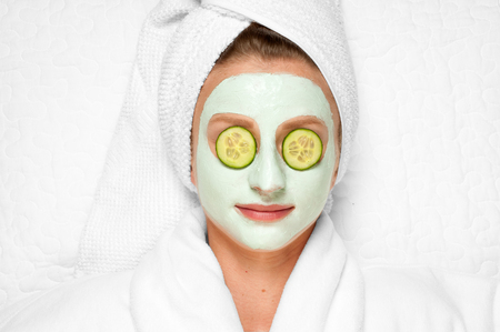 Beautiful young woman is getting facial clay mask at spa, lying with cucumbers on eyesの写真素材