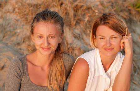 Two beautiful young girls sitting on sand at the beach look at the sunsetの写真素材