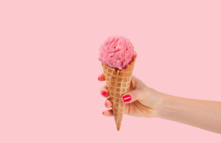 Hand holding strawberry ice cream cone on pink faded pastel color background. Strawberry ice cream in wafer cup.