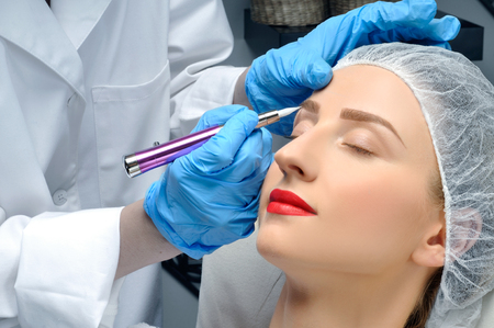 Microblading eyebrows. Cosmetologist making permanent makeup. Attractive woman getting facial care and tattoo at beauty salonの写真素材