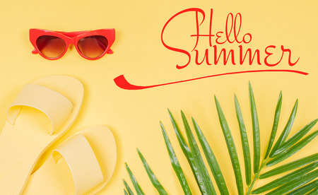 Hello Summer text on yellow background, sandals and sunglasses with green tropical palm leaf. Beach accessoriesの写真素材