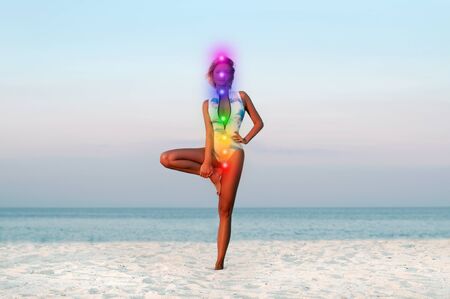 Woman is meditating with glowing seven chakras on the beach. Beautiful  woman is practicing yoga at sunset on sea shore. Kundalini meditation.の写真素材