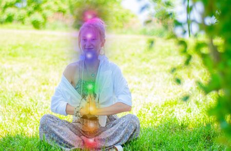 Happy woman is meditating in the lotus pose with glowing seven chakras on grass. Woman is practicing yoga on the park. Kundalini meditation.の写真素材