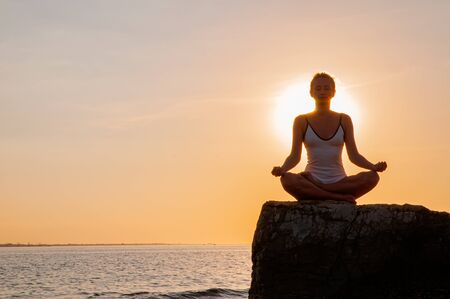 Woman is meditating on the calm beach at sunset. Woman is practicing yoga sitting on stone in Lotus pose at sunset. Silhouette of woman meditating on the beachの写真素材