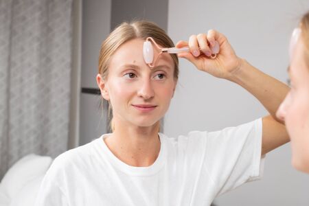 Beautiful woman is getting massage face using jade facial roller for skin care in front of the mirror at homeの写真素材