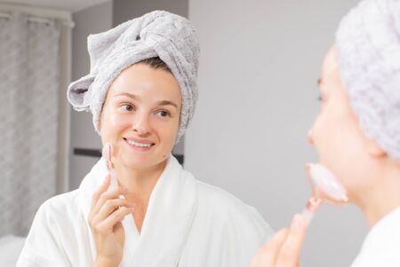 Beautiful woman is getting massage face using jade facial roller for skin care in front of the mirror at homeの写真素材