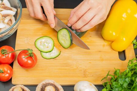 Woman is cutting cucumber on cut board on kitchen. Fresh vegetables for saladの写真素材
