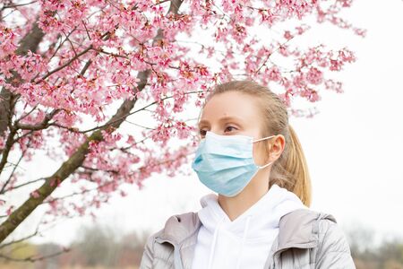 Beginning of Spring. Young woman in protective mask from pollen allergy, among blooming trees in park.の写真素材