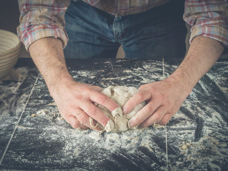 image shows two hands kneading dough for homemade sourdough bread or pizza; dark kitchen table with...