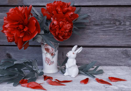 The bouquet of red peonies costs in a porcelain vase against the background of a wooden wall. The figure of a white porcelain rabbit is near.の写真素材