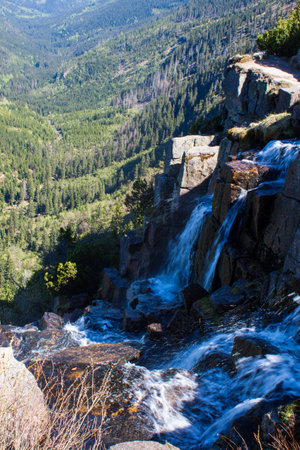 the water rushes down the rocks creating a waterfall. In the distance a mountainous landscape and coniferous forests with a clear sky.の写真素材
