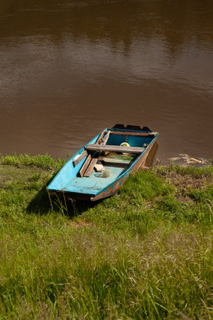 An old boat is in the grass by the river. There is sun, the water is brown.