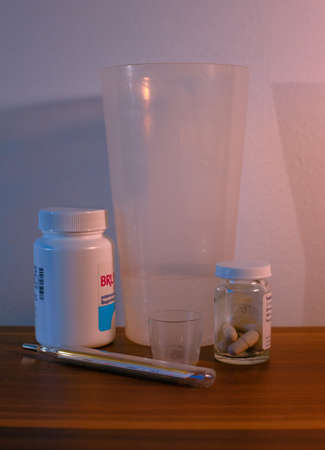 A thermometer, pills, tablets, a small measuring cup and a large plastic cup are gathered in front of a white wall.の写真素材