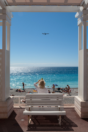 Promenade des anglais, Nice, France - September 2012 : Woman sitting on bench looking at the sea and a plane, framed by a white wooden arch.のeditorial素材