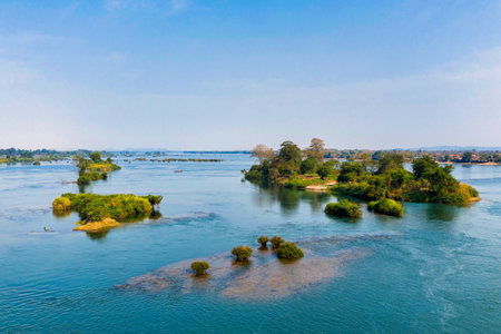 A broad stretch of the Mekong River flows past scattered green islets and a tree-lined riverbank in the 4000 Islands region of southern Laos. Clear daylight and calm water create a spacious riverscape with vibrant foliage and gentle reflections.の写真素材