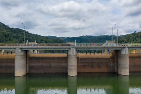 A modern concrete dam spans a calm reservoir, set against a background of densely forested hills and...