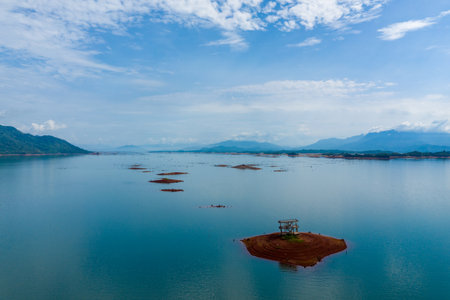 Aerial perspective of a small red earth islet with a lone structure surrounded by tranquil blue water in Nam Ngum Reservoir, Laos. Scattered islets and distant mountains create a sense of solitude and wide open space under a partly cloudy sky.の写真素材
