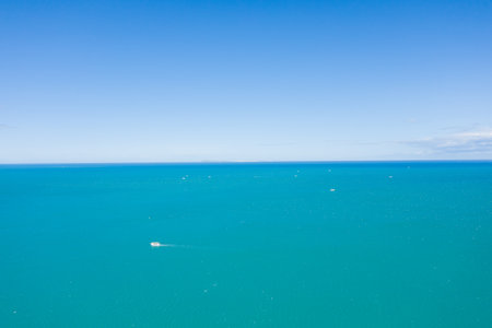 Minimalist aerial view of a single boat moving across vibrant turquoise water with a wide expanse of open sea and a cloudless blue sky, evoking freedom and tranquility.の写真素材