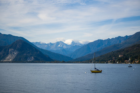 A yellow sailboat floats on the calm waters of Lake Maggiore in northern Italy, framed by forested hills and distant snow-capped alpine mountains beneath a sky streaked with clouds. The tranquil lakeside scene features subtle reflections and a peaceful atmosphere.の写真素材