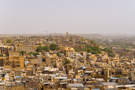 Expansive view of Jaisalmer with clustered golden sandstone buildings and scattered greenery under a hazy sky, capturing the texture and warmth of this historic desert city.の写真素材